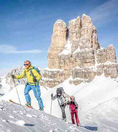 Due persone risalgono un pendio innevato con gli sci d’alpinismo, mentre imponenti cime dolomitiche si ergono sullo sfondo