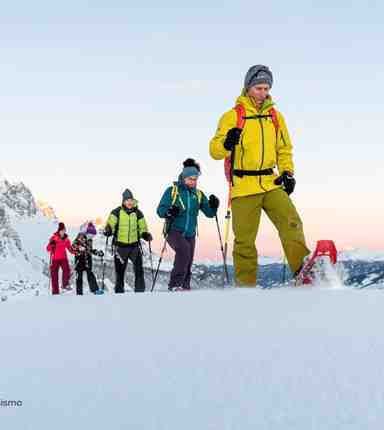 A group of people is snowshoeing through a snowy mountain landscape at sunset