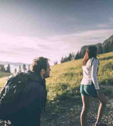 A man and a woman are hiking, surrounded by a wide alpine pasture landscape