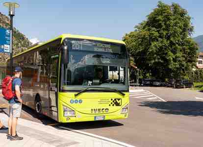 Three people are waiting at a bus stop for a green public bus in a sunny mountain town